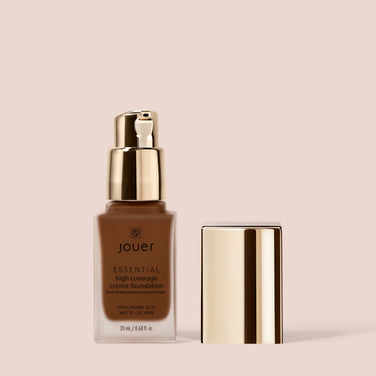  Image of a bottle of Jouer Cosmetics Essential Crème Foundation. The rectangular bottle features a gold pump dispenser with the cap placed beside it. This foundation, enriched with hyaluronic acid, offers a matte, oil-free finish and is both cruelty-free and paraben-free.