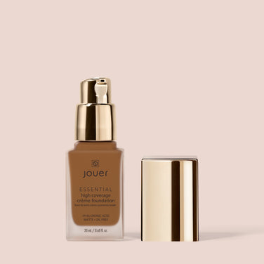  A bottle of Jouer Cosmetics Essential Crème Foundation is displayed against a light peach background. The glass bottle features a gold pump dispenser, with the cap removed and placed beside it. The label reads "hyaluronic acid, matte, oil free, gluten free, paraben free, cruelty free, 20 ml / 0.68 fl oz.