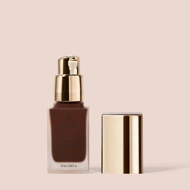  A bottle of Jouer Cosmetics Essential Crème Foundation in a frosted glass container with a gold pump dispenser and cap. Paraben Free and Cruelty Free, the cap is shown next to the bottle. The background is a light beige color.