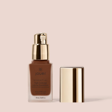  A bottle of Jouer Cosmetics Essential Crème Foundation, paraben-free and cruelty-free, with a gold pump dispenser and a removed gold cap is displayed against a beige background. The bottle contains 20ml/0.68 fl oz of foundation in a deep brown shade.