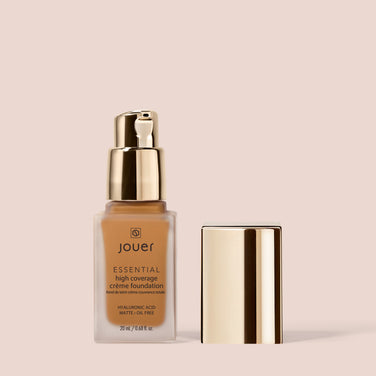  A bottle of Jouer Cosmetics Essential Crème Foundation is displayed on a beige background. The bottle, adorned with its name and product details, includes a gold pump dispenser. The cap, removed from the dispenser, stands upright beside the bottle. This foundation is both Cruelty Free and Paraben Free.