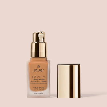  A bottle of Jouer Cosmetics' Essential Crème Foundation with a gold pump sits next to its shiny gold cap against a light beige background. The gluten-free, paraben-free, and cruelty-free formula holds 20ml (0.68 fl oz) of product and promotes a matte finish with hyaluronic acid.
