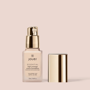  A bottle of Jouer Cosmetics Essential Crème Foundation with a gold pump dispenser sits next to its matching gold cap on a beige background. The 20 ml bottle boasts hyaluronic acid, a matte, oil-free finish, and proudly lists itself as cruelty-free and paraben-free.