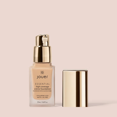  A bottle of Jouer Cosmetics' Essential Crème Foundation with a pump dispenser is shown. The square bottle and its cap are placed side by side. Labeled as containing hyaluronic acid, this foundation offers matte, oil-free coverage and is both paraben-free and cruelty-free.