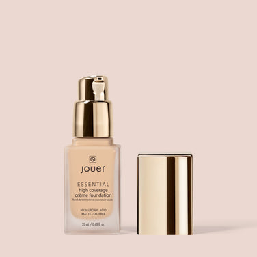  A bottle of the Essential Crème Foundation by Jouer Cosmetics, elegantly showcased on a neutral backdrop, reveals its beige liquid through a transparent design. The product features a gold pump dispenser with a matching cap placed next to it. This foundation is also Cruelty Free, providing beauty with a conscience.