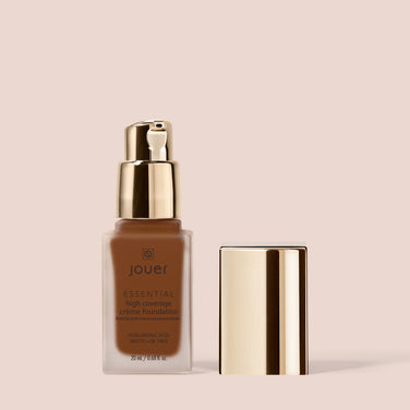  A bottle of Jouer Cosmetics Essential Crème Foundation with a gold pump dispenser is set against a beige background. The corresponding gold cap is positioned to the right of the bottle, which contains 20 ml (0.68 fl oz) of product. This formula is Paraben Free and Cruelty Free, making it a conscientious choice.