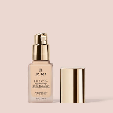  A bottle of Jouer Cosmetics Essential Crème Foundation, featuring a gold pump lid, is showcased next to its separated shiny gold cap against a beige background. The label highlights its content of hyaluronic acid, the provision of full coverage, and confirms it is paraben-free and cruelty-free.