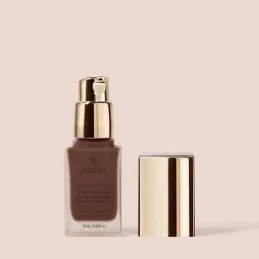  A rectangular bottle of Jouer Cosmetics Essential Crème Foundation with a gold pump dispenser and an accompanying gold cap placed next to it. The paraben-free bottle has a smooth, matte finish with black text. The background is a solid light beige color.