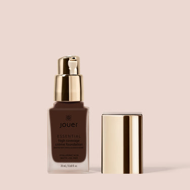  A bottle of Jouer Cosmetics Essential Crème Foundation. The elegant packaging is adorned with a gold pump dispenser and a matching gold cap placed beside it. The label indicates it contains 20 ml / 0.68 fl oz, highlights the presence of hyaluronic acid, and proudly states that it is Paraben Free. The background is beige.
