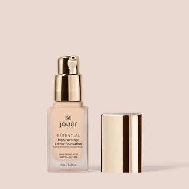  A bottle of Jouer Cosmetics' Essential Crème Foundation with a gold pump dispenser is displayed against a beige background. The cruelty-free product description in black text adorns the glass bottle, with a matching gold cap positioned beside it.