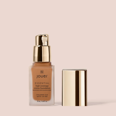  A bottle of Jouer Cosmetics Essential Crème Foundation with a gold pump dispenser is shown. The bottle is standing upright next to its matching gold cap, all set against a beige background. The text on the bottle highlights its high coverage, hyaluronic acid content, and matte, oil-free finish. It’s also cruelty free.