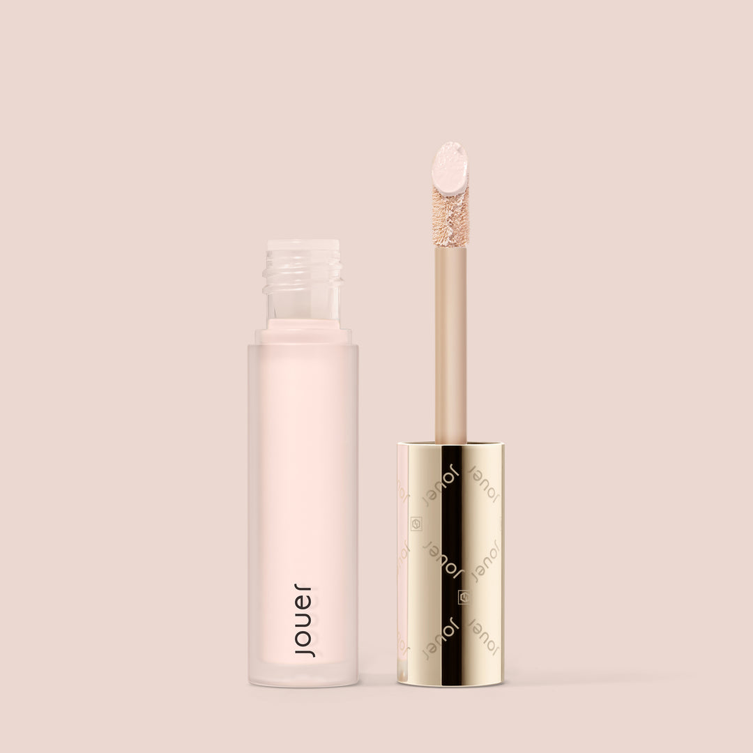 snow, A cylindrical tube of Essential Liquid Concealer from Jouer Cosmetics in a light pink hue, featuring a gold cap. The cap is removed to reveal a wand applicator with a doe-foot tip. The packaging and background are in soft beige tones, highlighting its paraben-free, vegan formula.