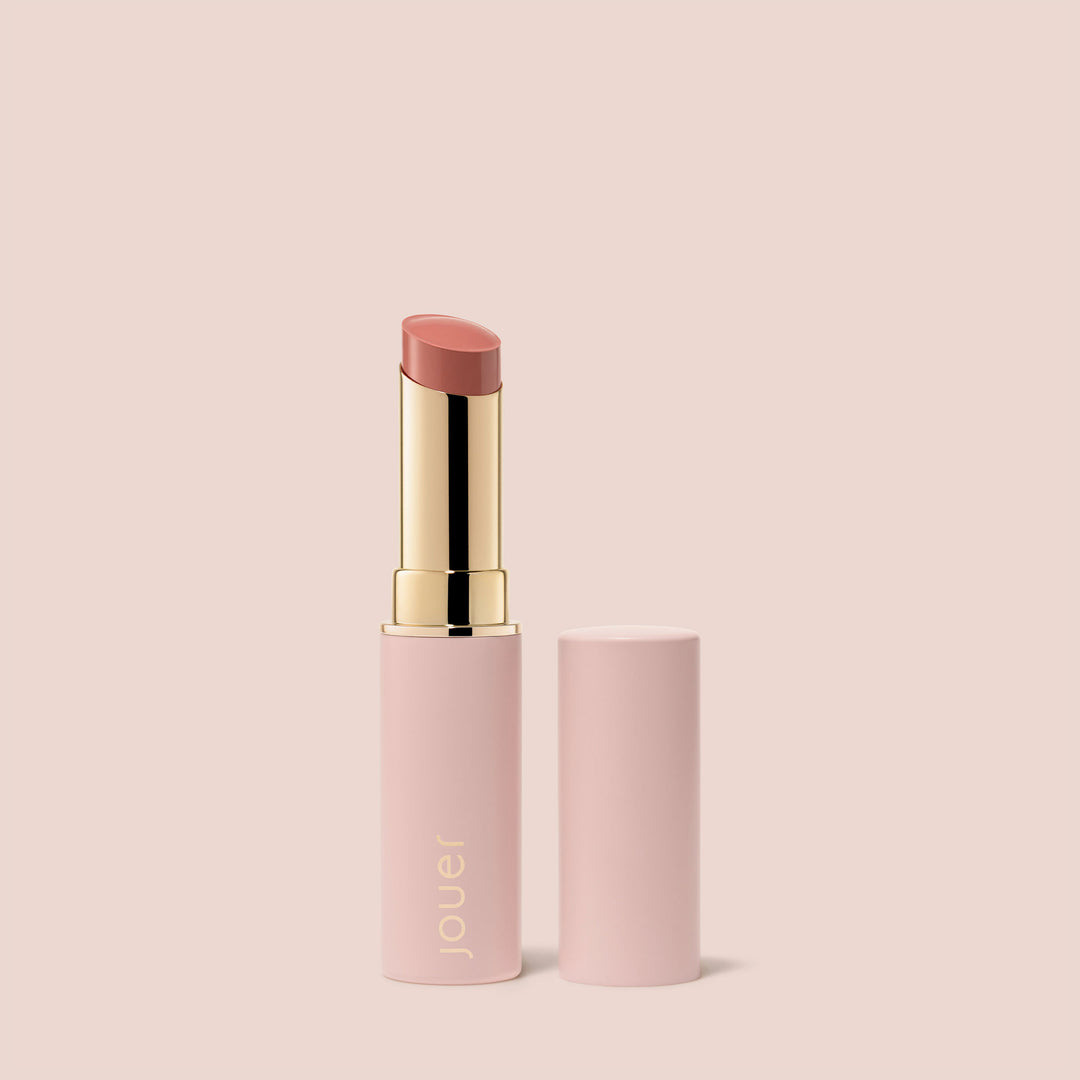 Sororite, A pink lipstick tube with a golden base and the cap removed, revealing a soft warm nude pink lipstick. The brand name "Jouer Cosmetics" is printed in gold on the body of the tube, which identifies this luxurious product as "Balm Bouche." The cap is placed next to the tube, all set against a light beige background. This exquisite product is also vegan and paraben-free.