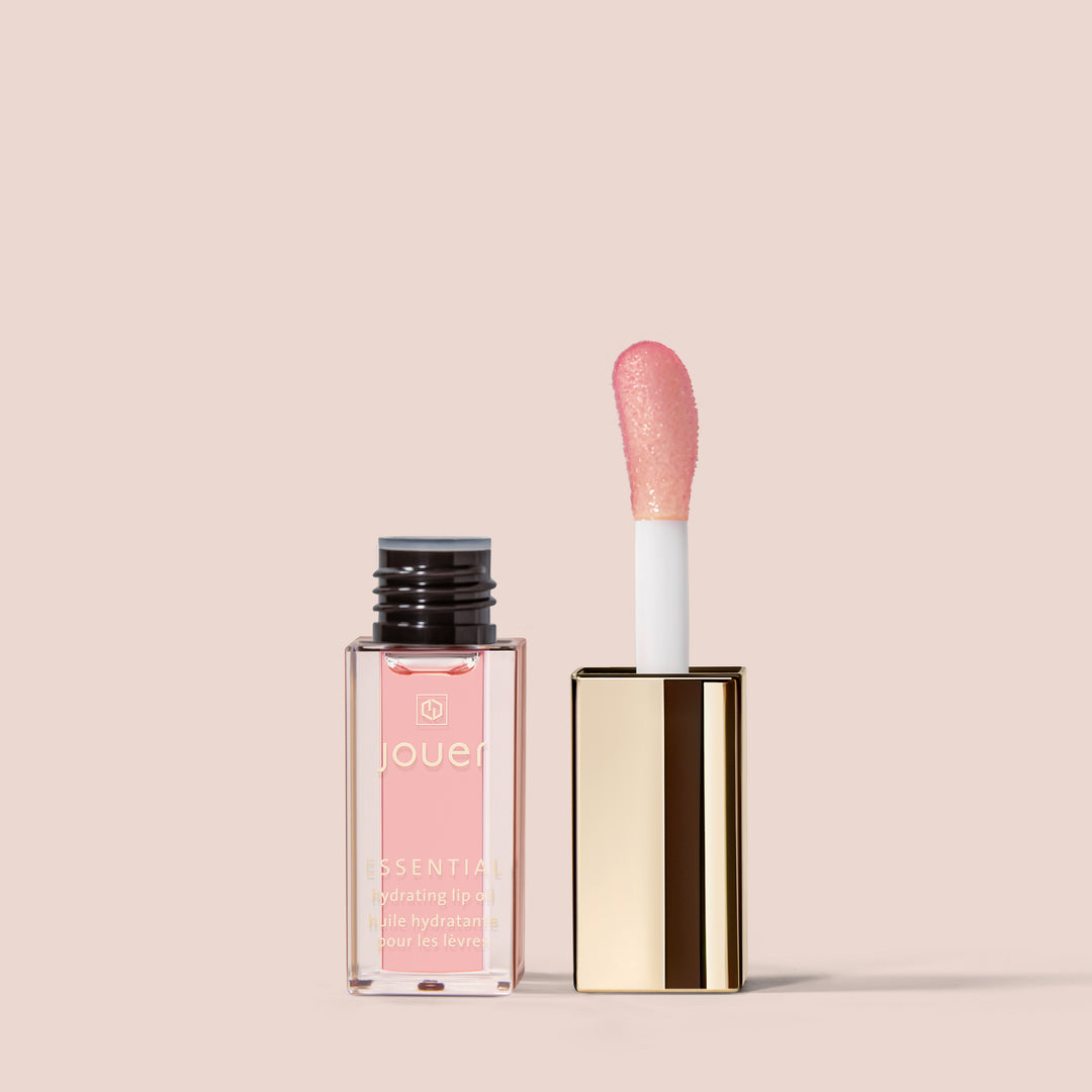 full size alt: A tube of Jouer Cosmetics' Essential Lip Oil, which is vegan and paraben-free, with its golden rectangular cap removed and placed upright beside it. The product's applicator, attached to the cap, features a pink-tinted sponge tip and stands against a light beige background.