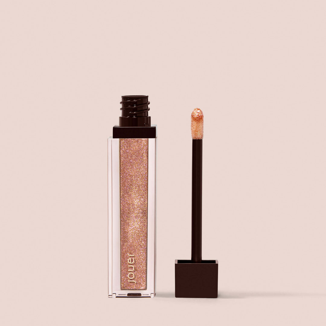 Rose Gold, A clear rectangular tube of glittery rose gold, paraben-free Lip Topper from Jouer Cosmetics with a black cap sits next to its applicator wand. The wand features a black handle and a sponge tip coated in the same vegan, glittery rose gold gloss, both set against a beige background.