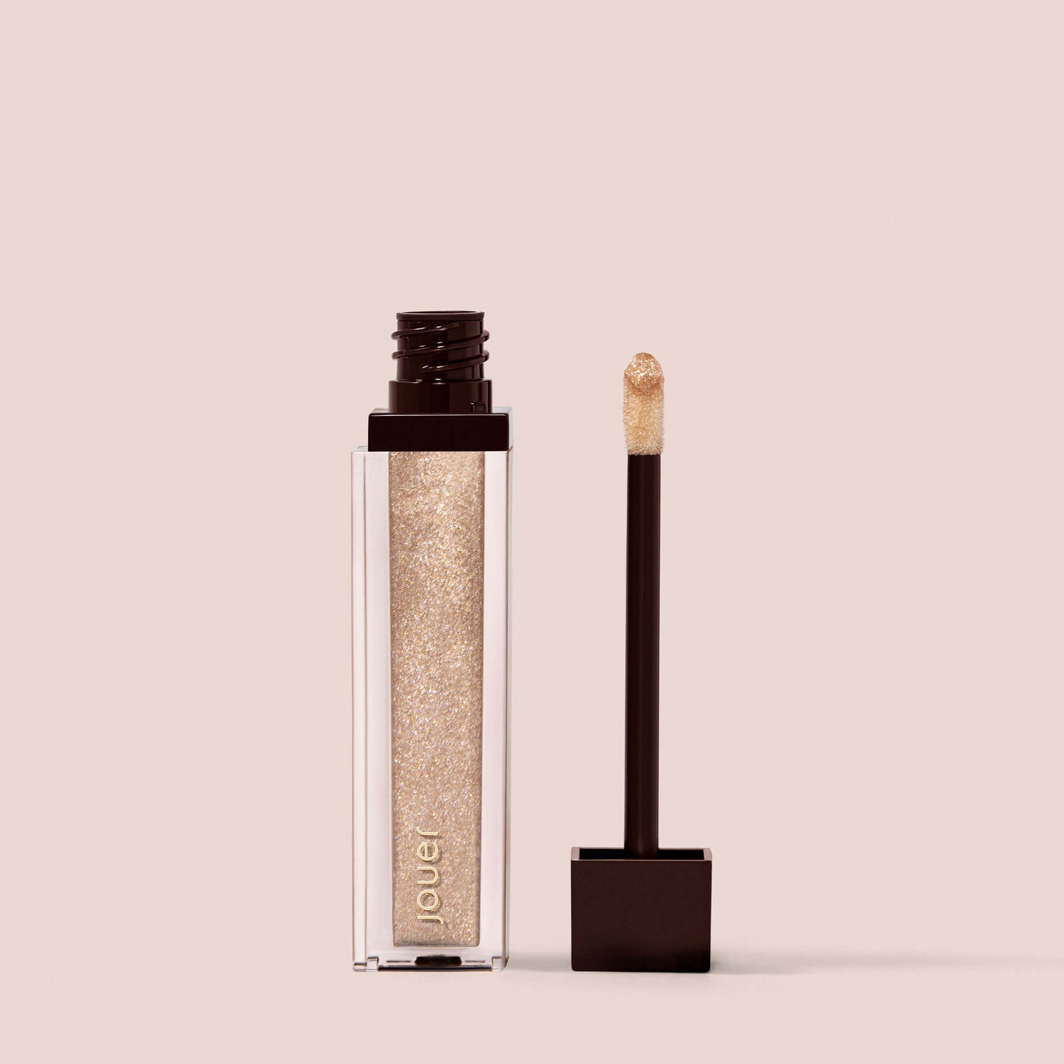 Skinny Dip, A tube of Jouer Cosmetics Lip Topper in a shimmering gold hue, paraben-free and vegan. The tube is open with the applicator wand resting beside it, displaying a bit of the gloss on its tip. The background is a plain beige color.