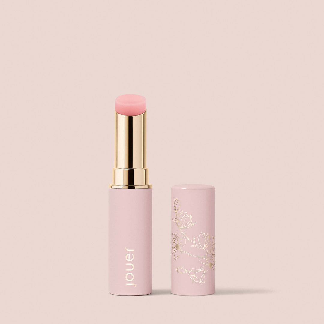 Naturel, An Essential Shine Balm from Jouer Cosmetics is displayed standing upright with its cap off and placed beside it. The cylindrical tube is light pink with "Jouer" written in white, while the cap features an elegant floral design in gold. This vegan and paraben-free product is set against a plain beige background.