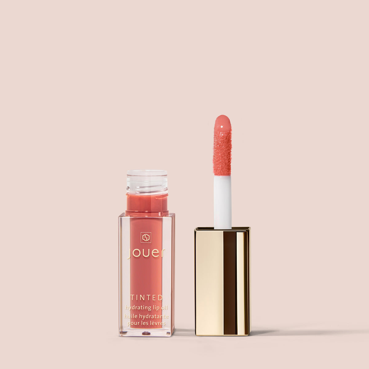 esprit, An open tube of Jouer Cosmetics Tinted Lip Oil, in a warm coral shade and formulated to be hydrating and paraben-free, sits with its applicator wand resting beside it. The lip oil is elegantly presented in a rectangular, transparent container topped with a shiny gold lid. The background is a soft peachy beige color.