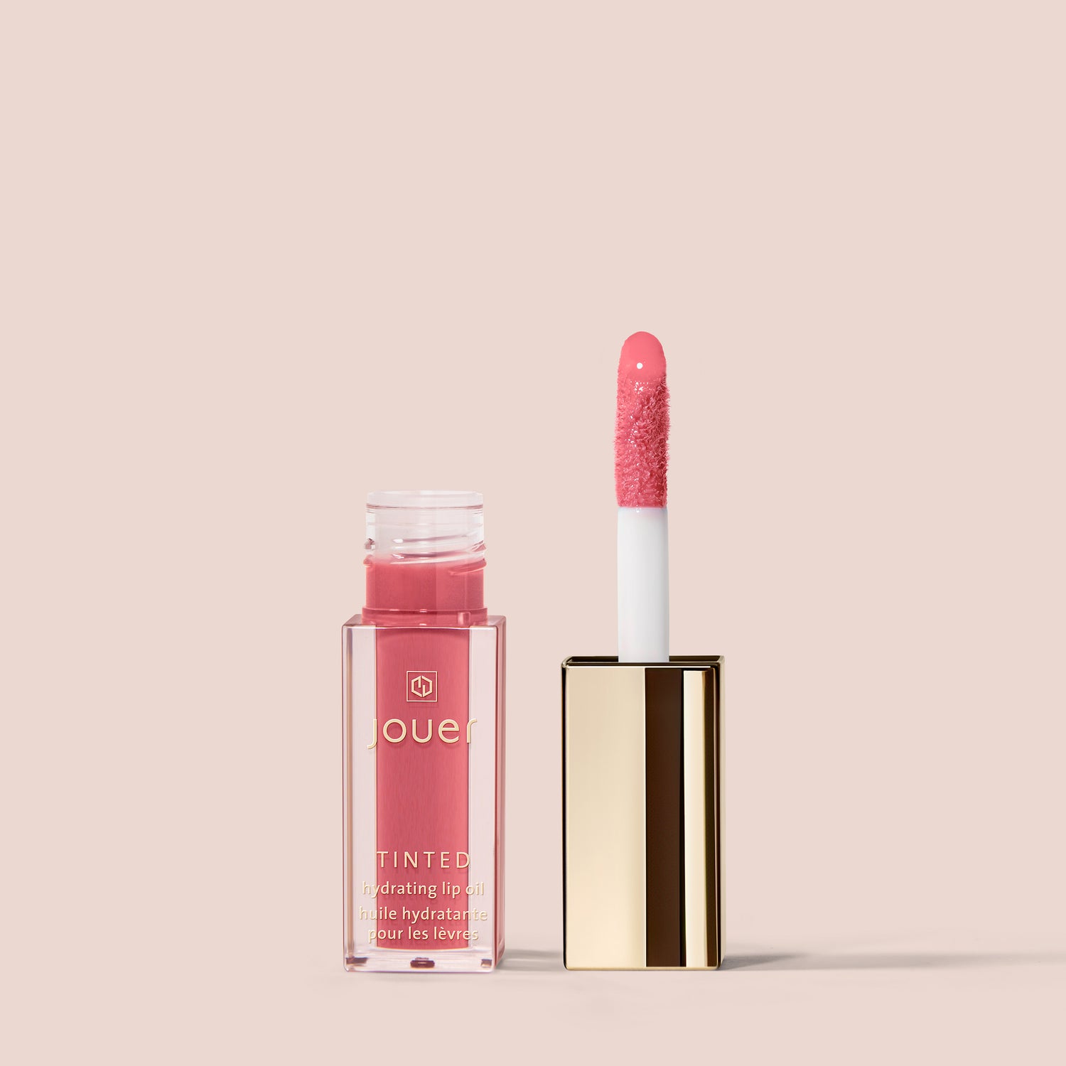 reve, A tube of Jouer Cosmetics Tinted Lip Oil with gold accents is displayed against a beige background. The opened tube reveals the pink-hued applicator wand standing upright next to the container. The label reads, "Jouer Tinted, Hydrating Lip Oil." This vegan and cruelty-free formula offers a paraben-free beauty experience.